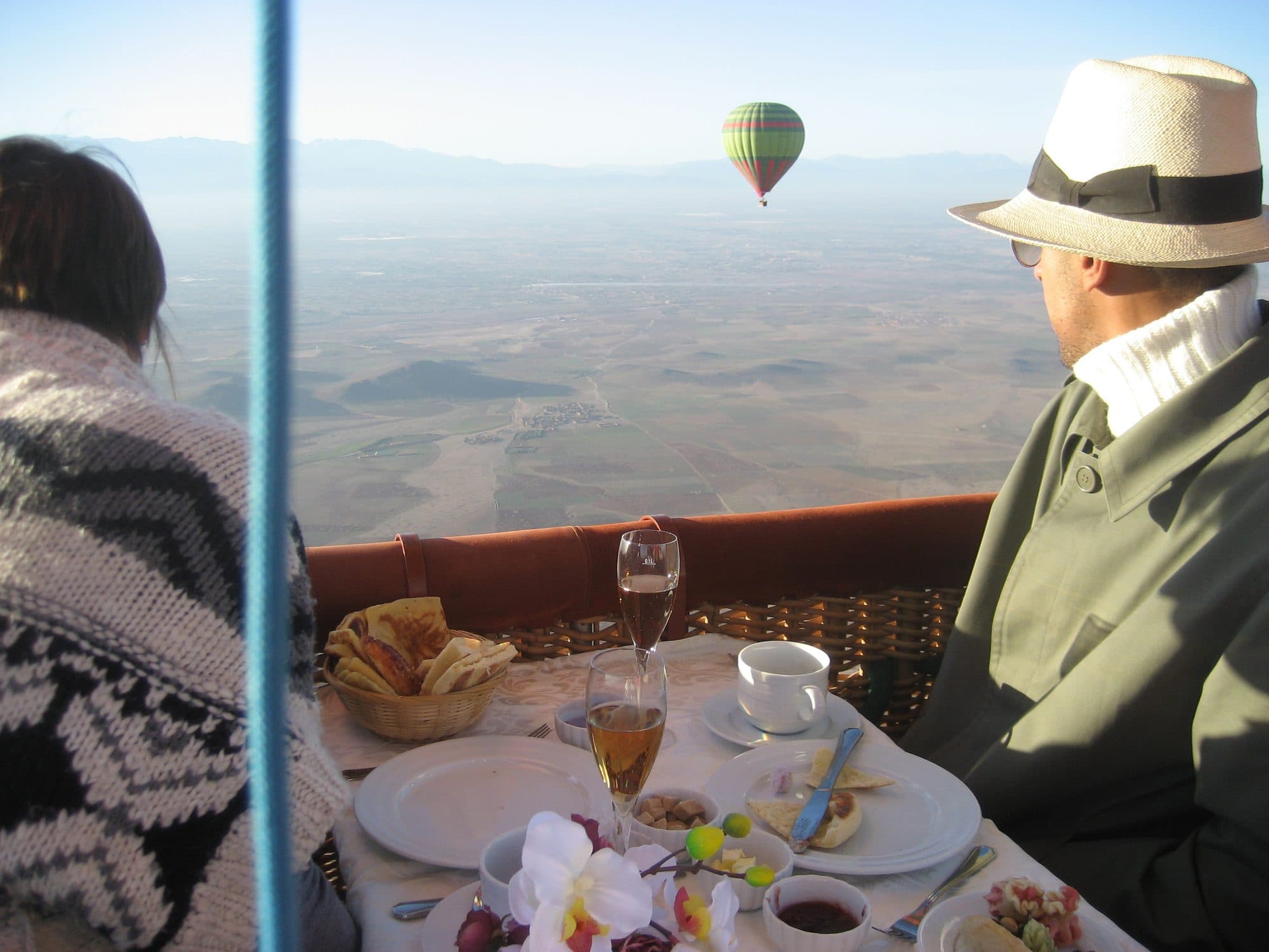 Royal breakfast table set inside a hot air balloon basket over the Marrakech countryside, with the Atlas Mountains on the horizon.